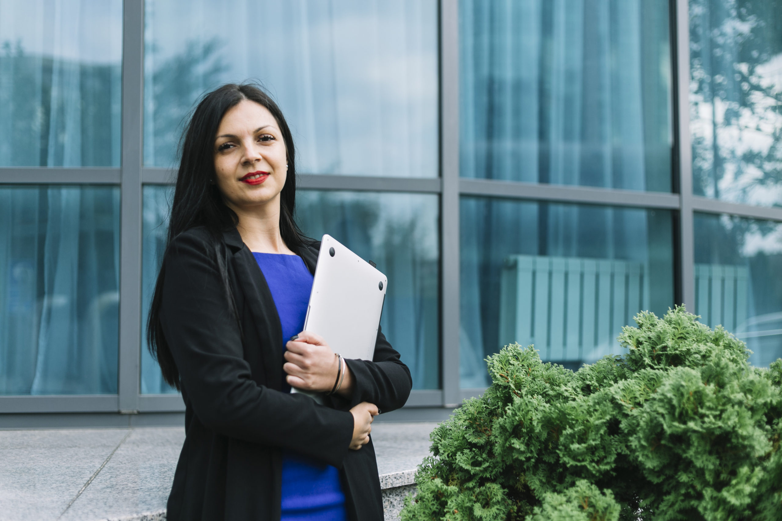 portrait-smiling-businesswoman-holding-laptop-front-glass-building-scaled